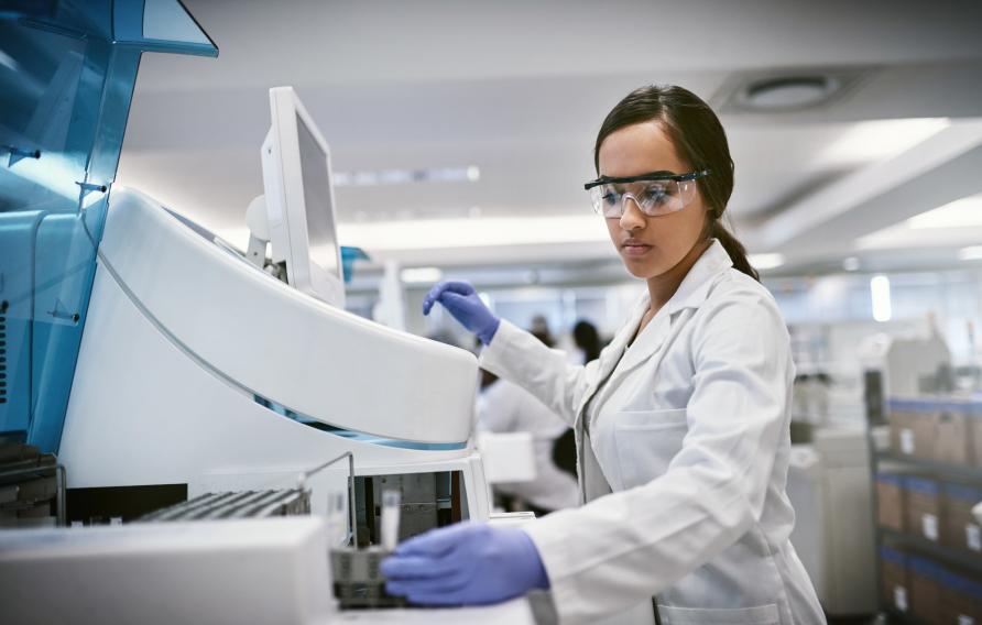 Medium skinned woman with dark hair in a lab coat, safety glasses and blue gloves works in a lab environment